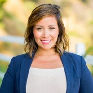 Person looking directly forward, smiling, wearing a dark blue blazer and white blouse.