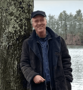 Photograph of George Kinder, standing next to tree, wearing casual jacket and hat