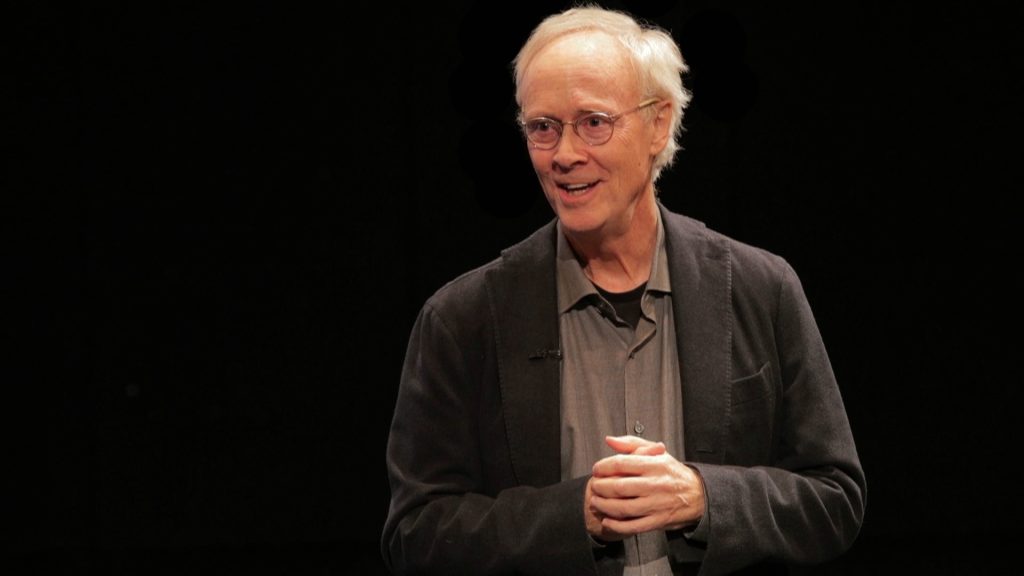 Man in a gray sports jacket and shirt speaking with a black background.