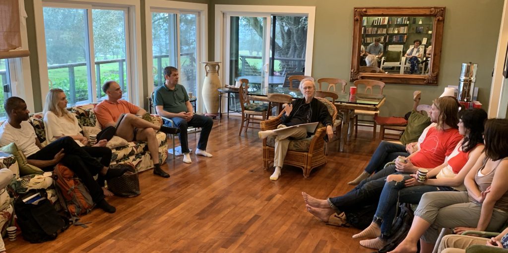 A dozen participants in casual dress sit in a living room listening to a teacher.