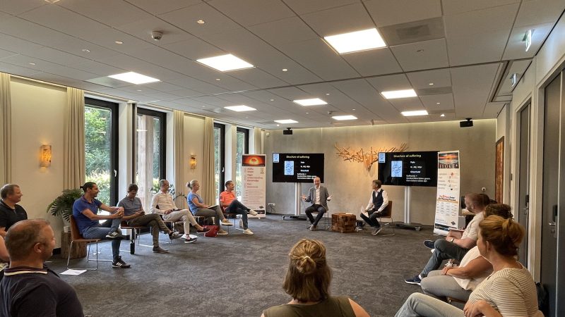 Group of 14 adults sitting in a circle indoors, wearing casual clothes. Two of the adults are leading the others in a lively discussion.